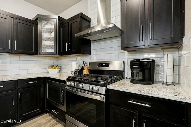 kitchen with light stone counters, dark cabinetry, wall chimney range hood, stainless steel gas stove, and tasteful backsplash