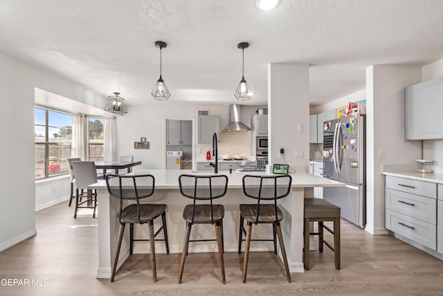 kitchen with light wood-style flooring, wall chimney exhaust hood, appliances with stainless steel finishes, a kitchen breakfast bar, and light countertops