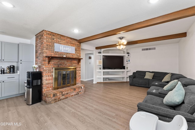 living area featuring visible vents, light wood-style flooring, beamed ceiling, a textured ceiling, and a brick fireplace