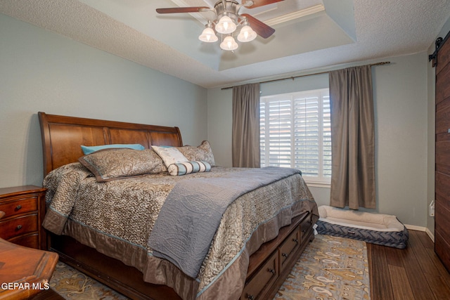 bedroom with a barn door, ceiling fan, wood finished floors, a tray ceiling, and a textured ceiling