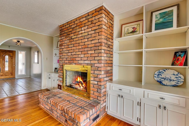 living room featuring light wood-style flooring, arched walkways, ornamental molding, a textured ceiling, and a brick fireplace