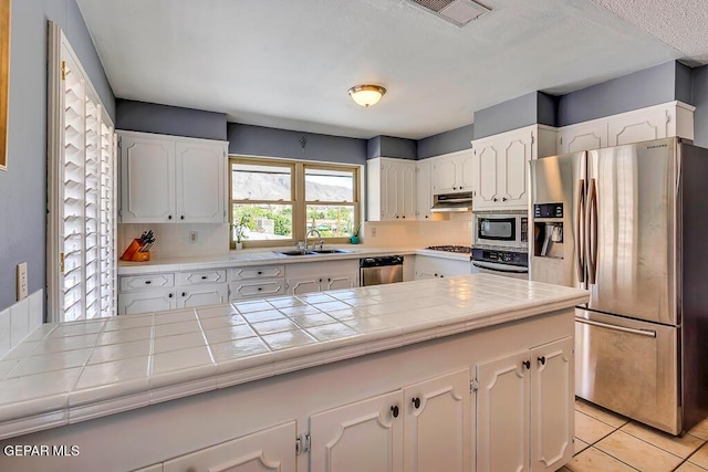 kitchen with a sink, tile counters, white cabinets, under cabinet range hood, and appliances with stainless steel finishes