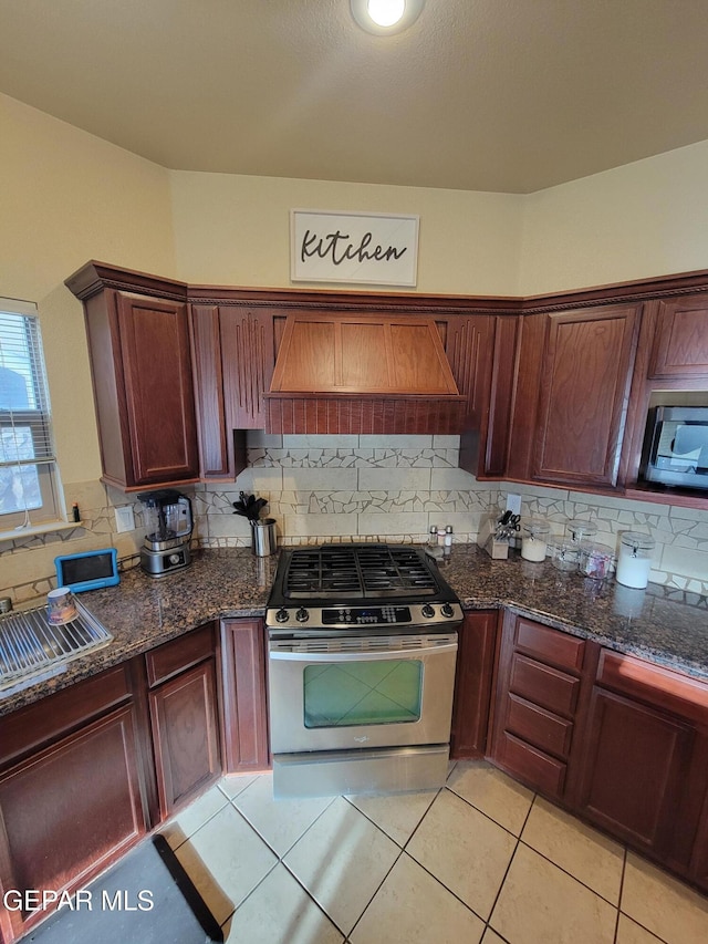 kitchen featuring light tile patterned flooring, appliances with stainless steel finishes, range hood, tasteful backsplash, and dark stone countertops