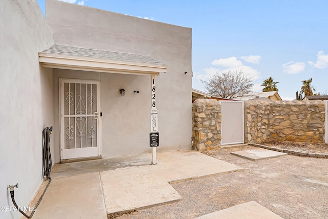 entrance to property with roof with shingles, a patio area, fence, and stucco siding