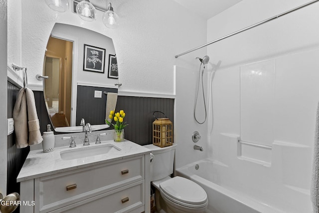 bathroom with toilet, wainscoting, shower / bathtub combination, a textured wall, and vanity