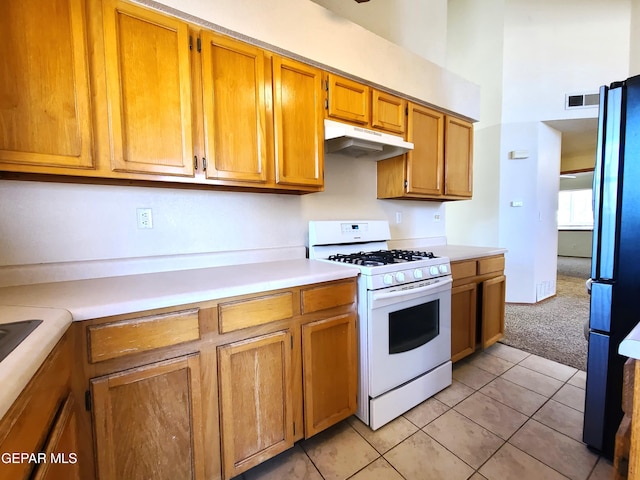 kitchen featuring white gas stove, under cabinet range hood, freestanding refrigerator, brown cabinetry, and light countertops