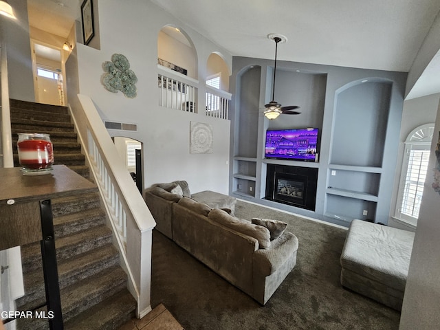 living room with visible vents, built in features, stairway, a glass covered fireplace, and a ceiling fan