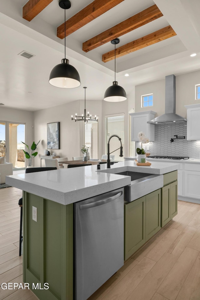 kitchen featuring an island with sink, a sink, stainless steel dishwasher, green cabinets, and wall chimney range hood