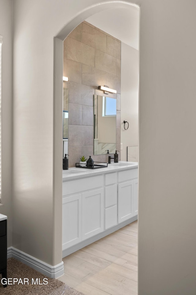 full bathroom with wood finished floors, baseboards, double vanity, a sink, and decorative backsplash