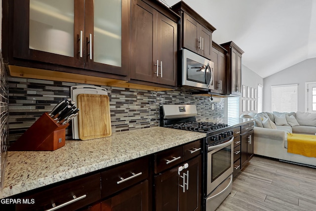 kitchen featuring dark brown cabinetry, appliances with stainless steel finishes, glass insert cabinets, light stone countertops, and vaulted ceiling