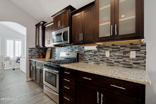 kitchen featuring light wood finished floors, backsplash, dark brown cabinetry, arched walkways, and stainless steel appliances