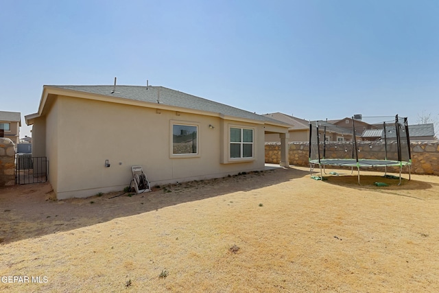 rear view of house with stucco siding, a trampoline, roof with shingles, and fence