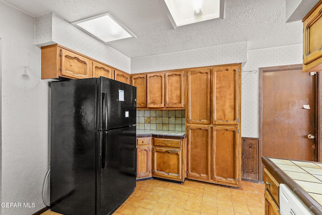 kitchen with a textured ceiling, freestanding refrigerator, tile countertops, brown cabinetry, and wallpapered walls