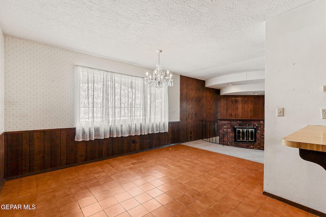 unfurnished living room with a textured ceiling, an inviting chandelier, wood walls, wainscoting, and a brick fireplace