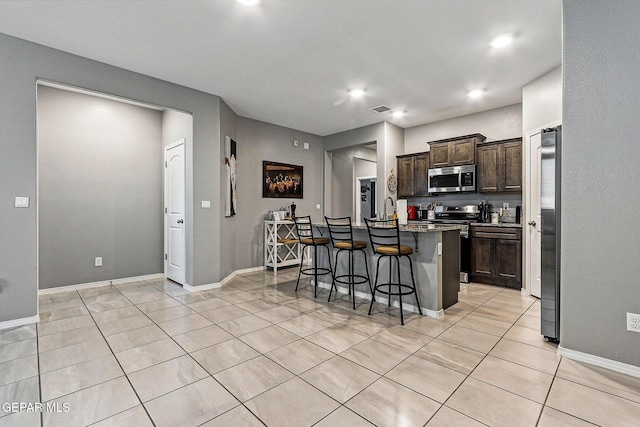 kitchen featuring a kitchen island with sink, a kitchen breakfast bar, stainless steel appliances, dark brown cabinetry, and light tile patterned flooring