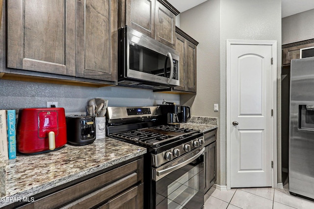 kitchen featuring dark brown cabinetry, a textured wall, light tile patterned flooring, stone countertops, and stainless steel appliances