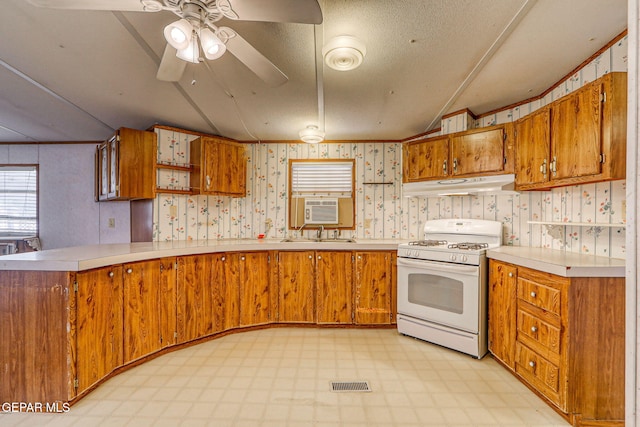 kitchen with wallpapered walls, under cabinet range hood, light floors, white gas range oven, and brown cabinets