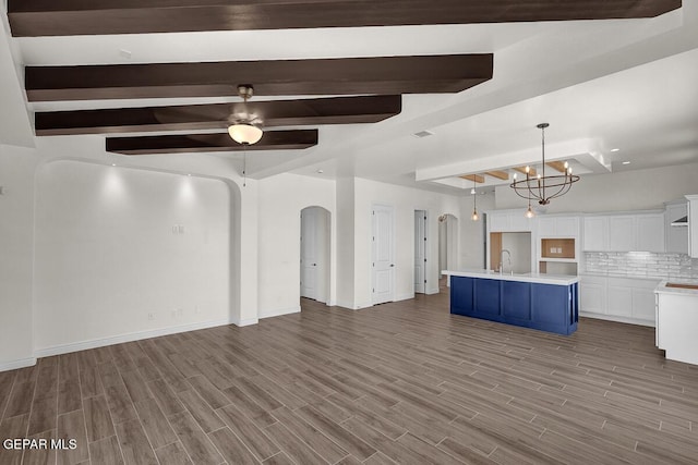 unfurnished living room featuring wood finish floors, beamed ceiling, ceiling fan with notable chandelier, a sink, and arched walkways