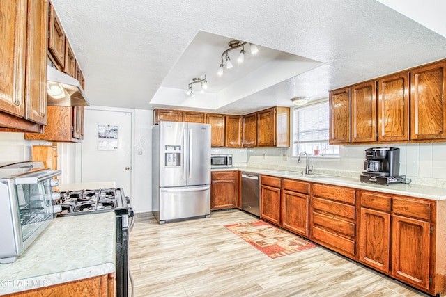 kitchen featuring under cabinet range hood, brown cabinets, stainless steel appliances, a raised ceiling, and a sink