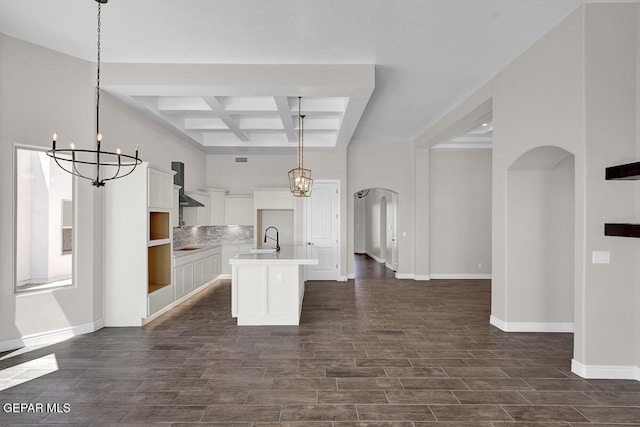 kitchen with coffered ceiling, an inviting chandelier, arched walkways, wall chimney range hood, and backsplash