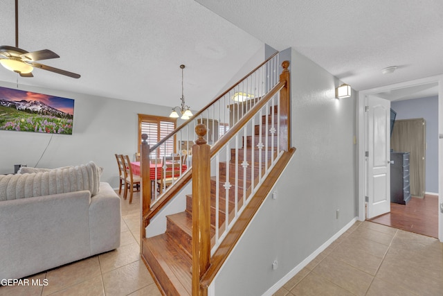 staircase featuring baseboards, a textured ceiling, tile patterned flooring, and ceiling fan with notable chandelier