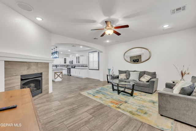 living area featuring a ceiling fan, visible vents, arched walkways, light wood-style floors, and a tiled fireplace