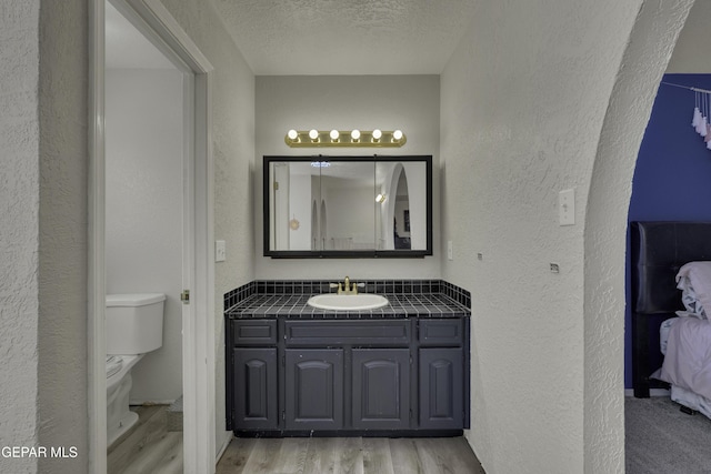 bathroom with toilet, a textured ceiling, wood finished floors, vanity, and a textured wall