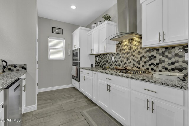 kitchen with white cabinetry, stainless steel appliances, wall chimney range hood, decorative backsplash, and light stone countertops