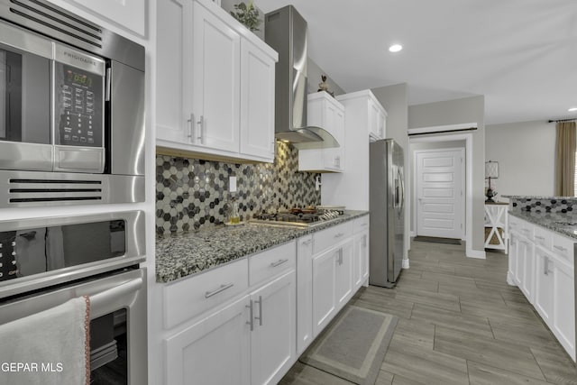 kitchen with wall chimney range hood, light stone counters, decorative backsplash, white cabinets, and stainless steel appliances