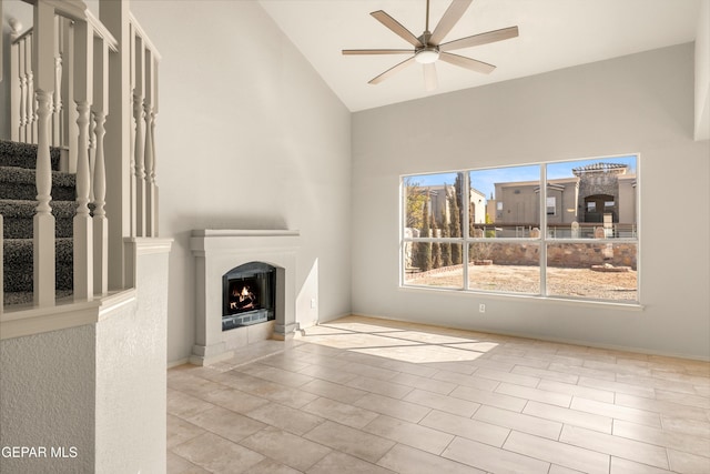 unfurnished living room featuring stairs, high vaulted ceiling, a warm lit fireplace, and a ceiling fan