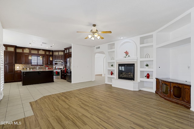 unfurnished living room with light wood finished floors, ceiling fan, built in features, arched walkways, and a glass covered fireplace