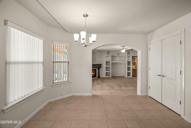 unfurnished dining area featuring tile patterned flooring, baseboards, a lit fireplace, ceiling fan with notable chandelier, and arched walkways