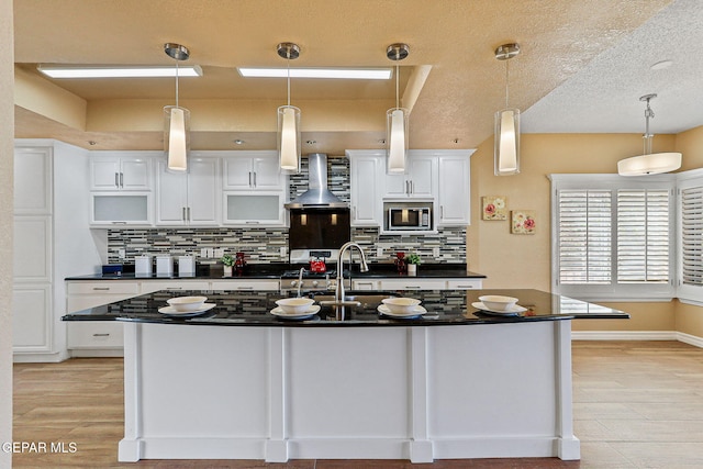 kitchen featuring stainless steel microwave, dark countertops, wall chimney exhaust hood, and white cabinetry