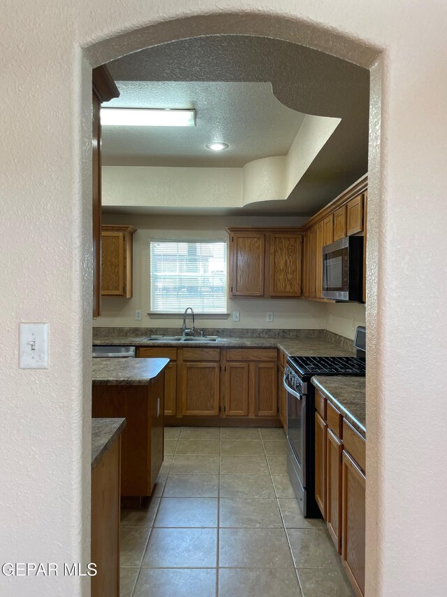 kitchen with light tile patterned flooring, dark countertops, a sink, brown cabinetry, and stainless steel appliances