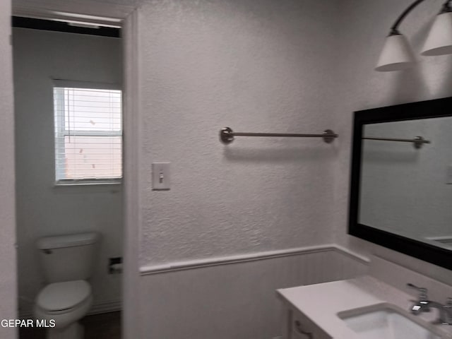 bathroom with a textured wall, toilet, vanity, and wainscoting