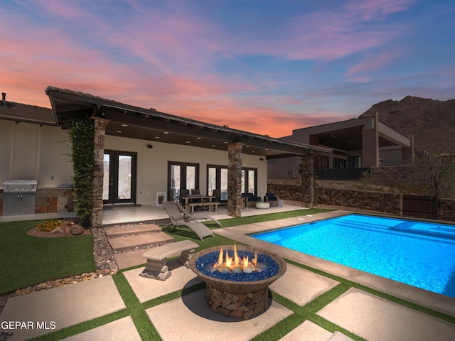 pool at dusk with an outdoor fire pit, area for grilling, a fenced in pool, a patio area, and french doors