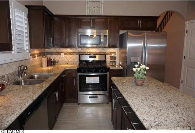 kitchen with appliances with stainless steel finishes, a sink, dark brown cabinets, light stone counters, and tasteful backsplash