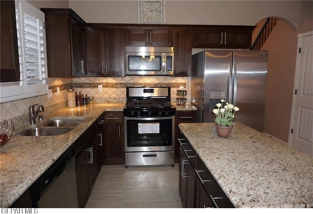 kitchen with appliances with stainless steel finishes, a sink, light stone counters, dark brown cabinetry, and backsplash
