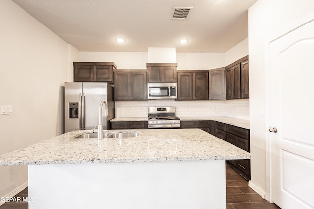 kitchen with stainless steel appliances, a sink, dark brown cabinetry, a kitchen island with sink, and tasteful backsplash