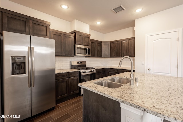 kitchen featuring appliances with stainless steel finishes, a sink, dark brown cabinets, tasteful backsplash, and light stone counters