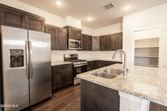 kitchen with appliances with stainless steel finishes, a sink, dark brown cabinets, light stone counters, and backsplash