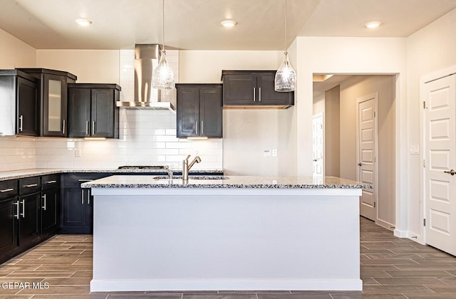 kitchen featuring light stone countertops, decorative backsplash, a center island with sink, wall chimney range hood, and wood finish floors