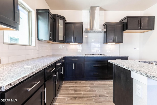 kitchen featuring glass insert cabinets, stainless steel gas stovetop, wall chimney range hood, backsplash, and wood tiled floor