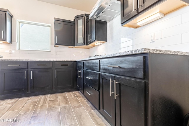 kitchen featuring wall chimney range hood, light wood-type flooring, decorative backsplash, and light stone countertops
