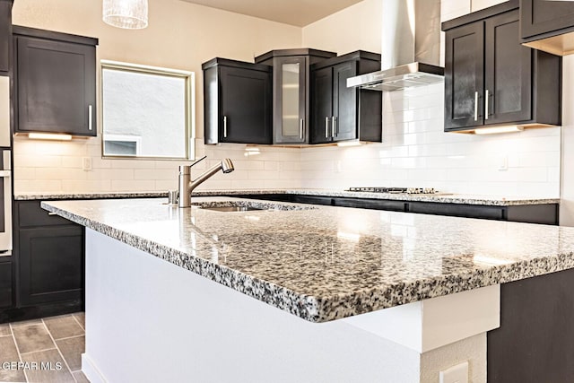 kitchen with light stone counters, decorative backsplash, stainless steel appliances, a sink, and wall chimney exhaust hood
