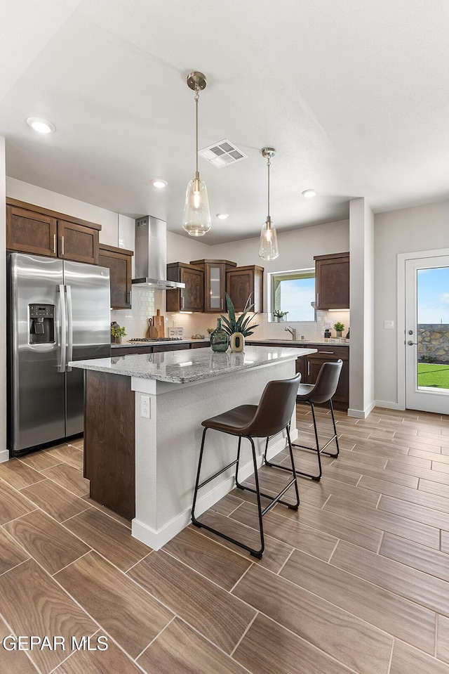 kitchen featuring visible vents, a center island, stainless steel refrigerator with ice dispenser, wall chimney exhaust hood, and dark brown cabinetry
