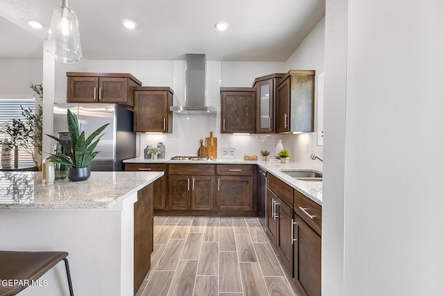 kitchen featuring wall chimney range hood, dark brown cabinets, appliances with stainless steel finishes, a sink, and hanging light fixtures