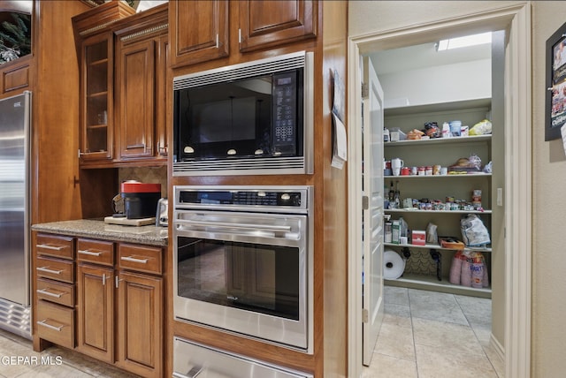 kitchen with a warming drawer, glass insert cabinets, light stone countertops, brown cabinetry, and appliances with stainless steel finishes