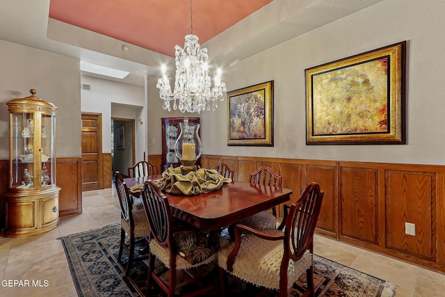 dining space with wooden walls, a notable chandelier, wainscoting, a raised ceiling, and a skylight