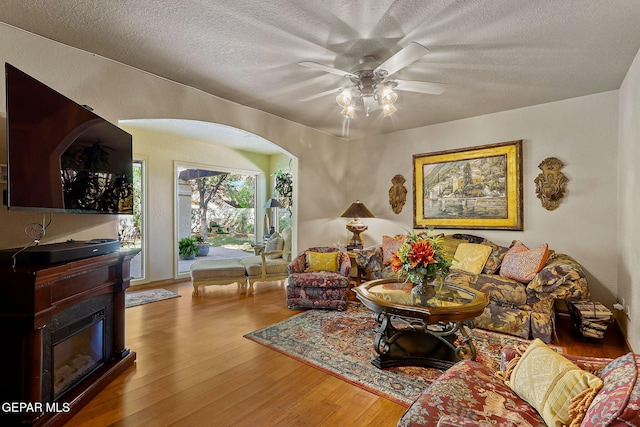 living room with a ceiling fan, a textured ceiling, arched walkways, wood finished floors, and a glass covered fireplace